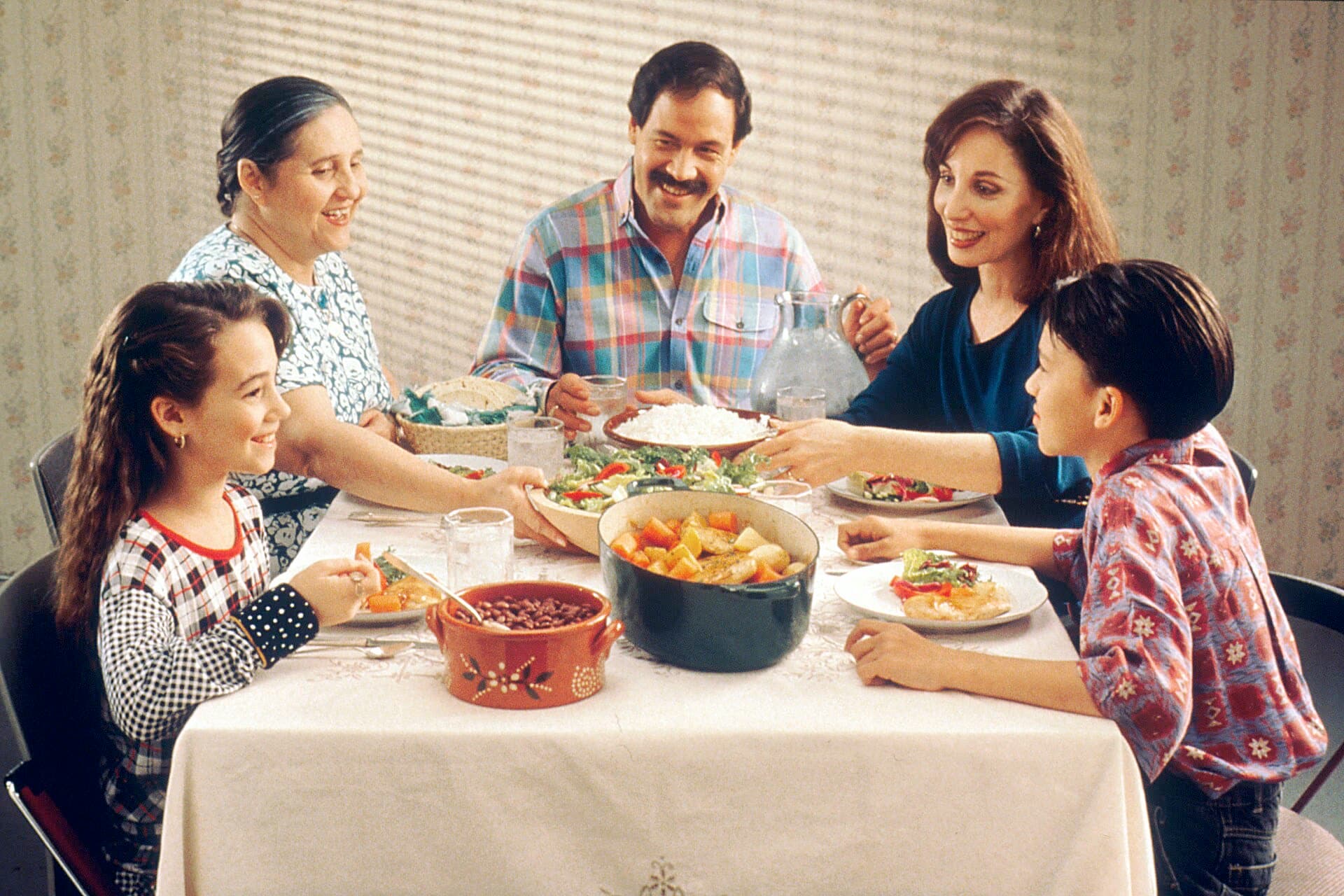 A family of five across three generations at a dinner table with a white cloth, passing a bowl of rice and a pot of vegetables between them and smiling.