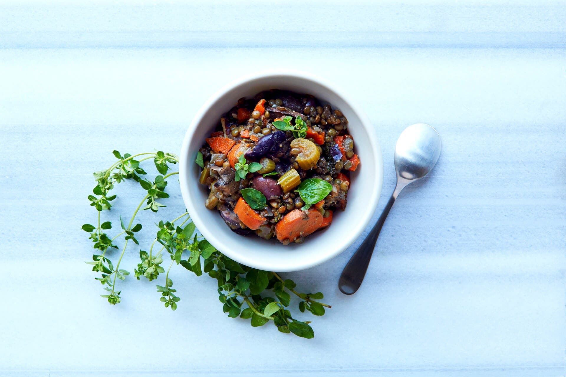 A white bowl of cooked green lentils with carrot, celery, olives and fresh basil, a spoon beside it on a pale table.