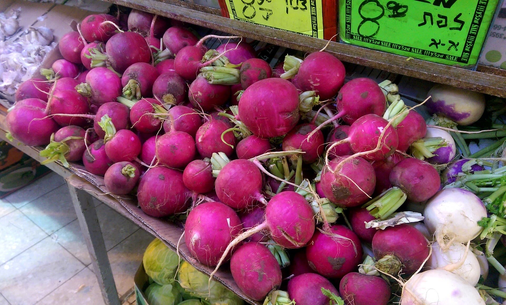A crate of bright pink radishes with their green tops, beside turnips and cabbages, under handwritten Hebrew price signs at a market stall in Jerusalem.