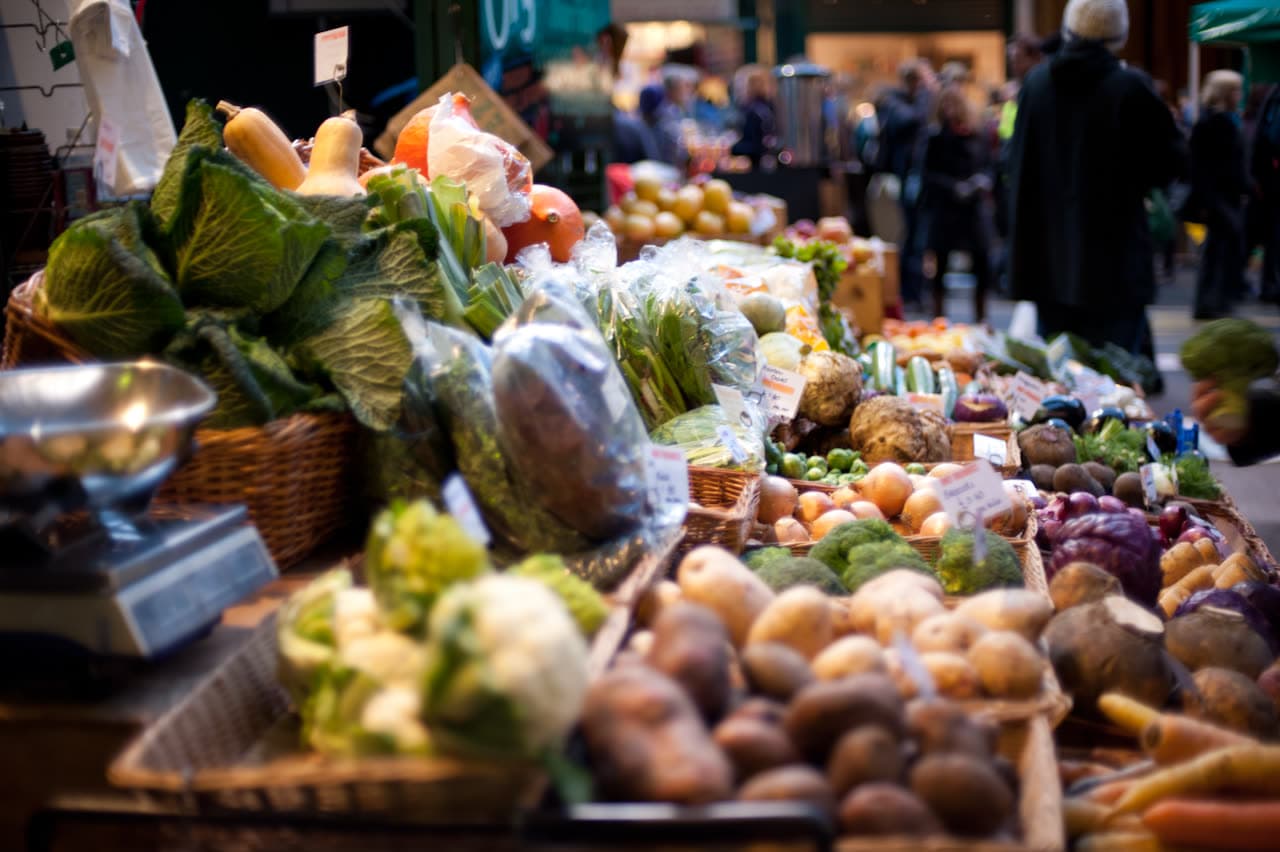A market stall piled with vegetables in baskets: cabbages, squash, leeks, potatoes, onions, broccoli, red cabbage and carrots, with shoppers blurred behind.